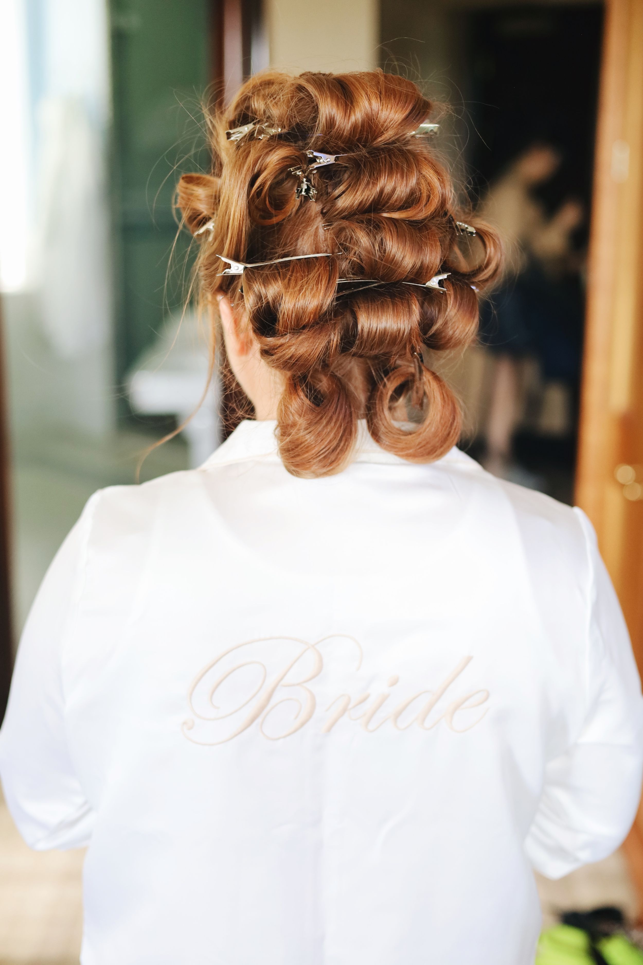 Back view of auburn hair in pin curls with 'Bride' text on white robe during wedding preparations.