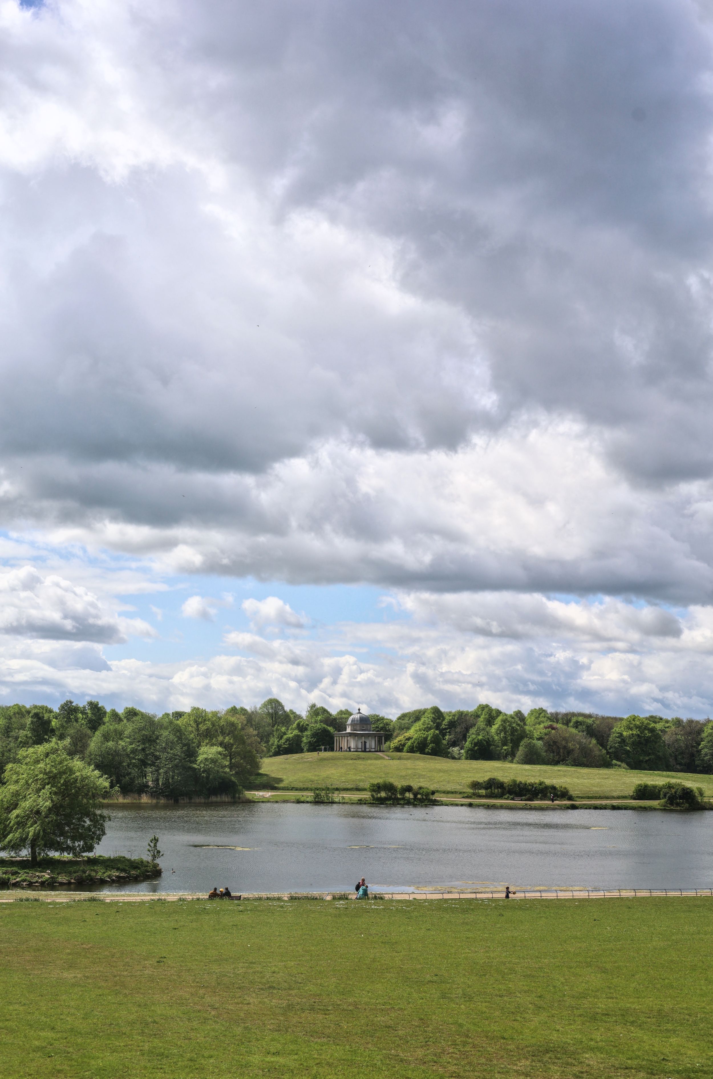 Scenic lakeside landscape with dramatic cloudy sky over green fields and trees.