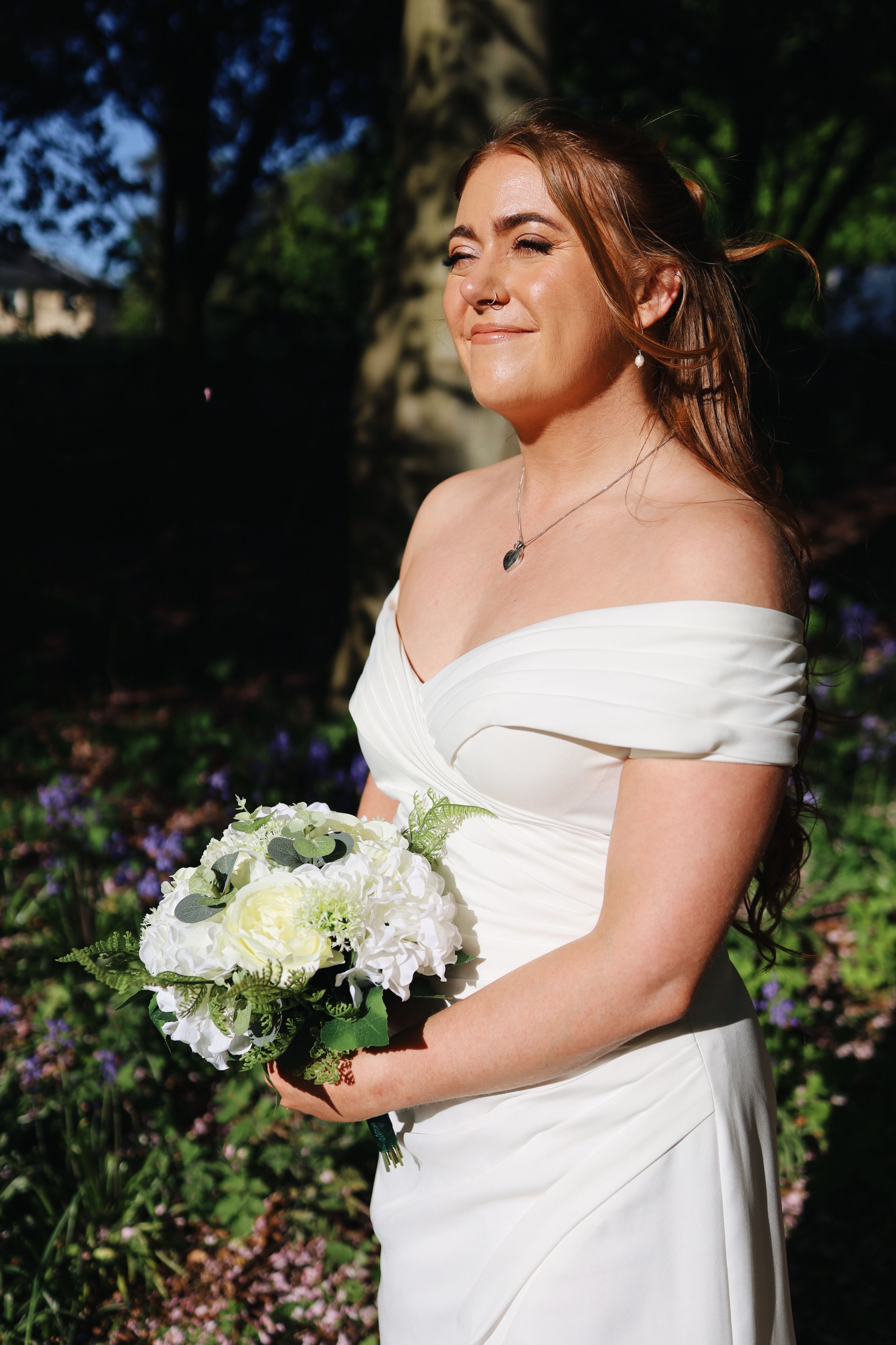 Bride in off-shoulder white dress holding white and green bouquet in garden setting.