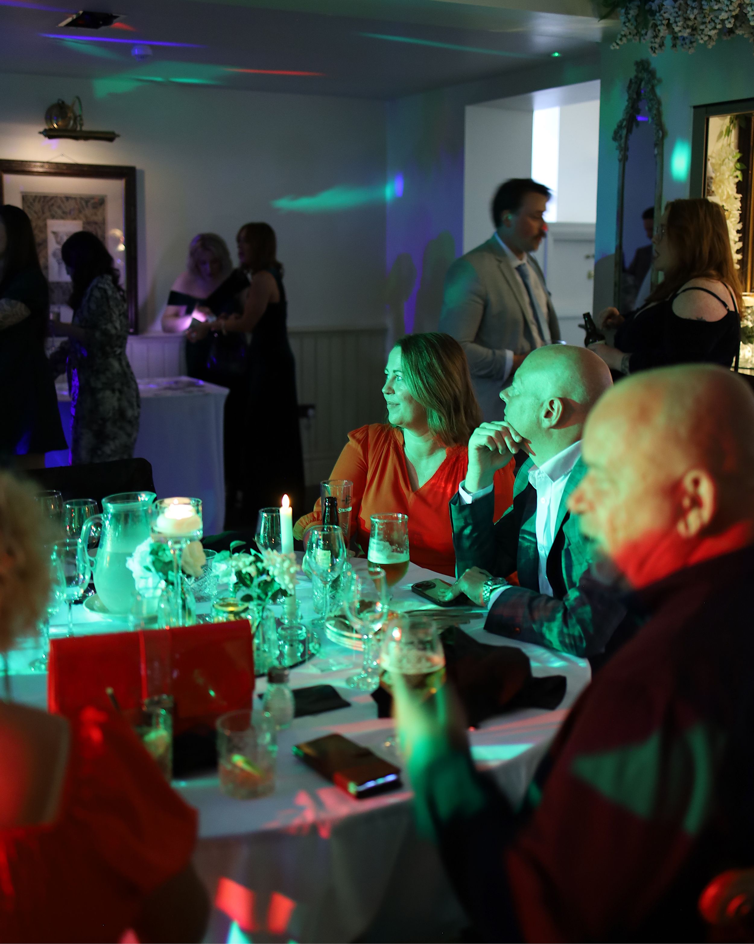 Reception guests sit at decorated tables illuminated by colorful ambient lighting.