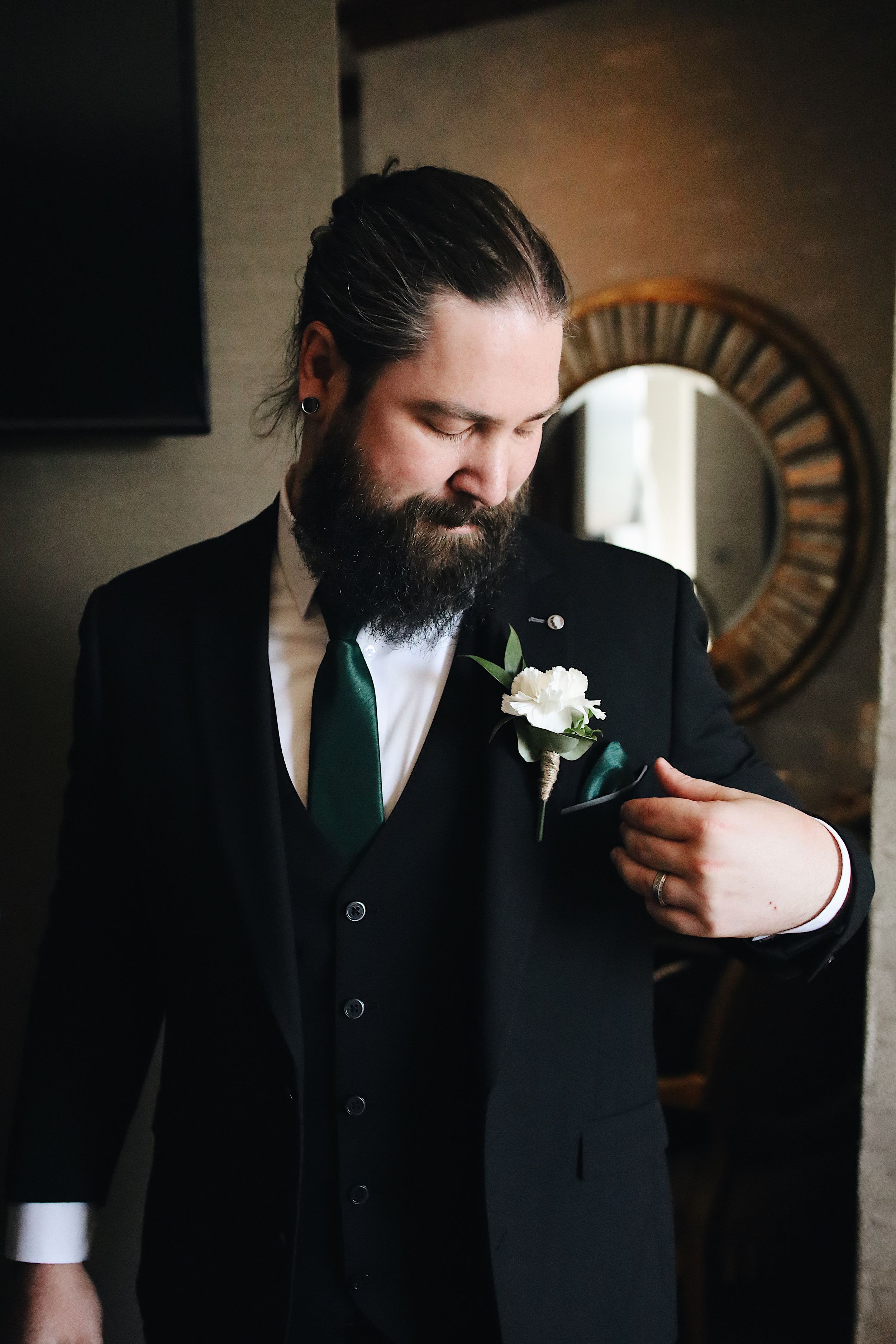 A bearded man in a black suit with green tie smells a white boutonniere flower.