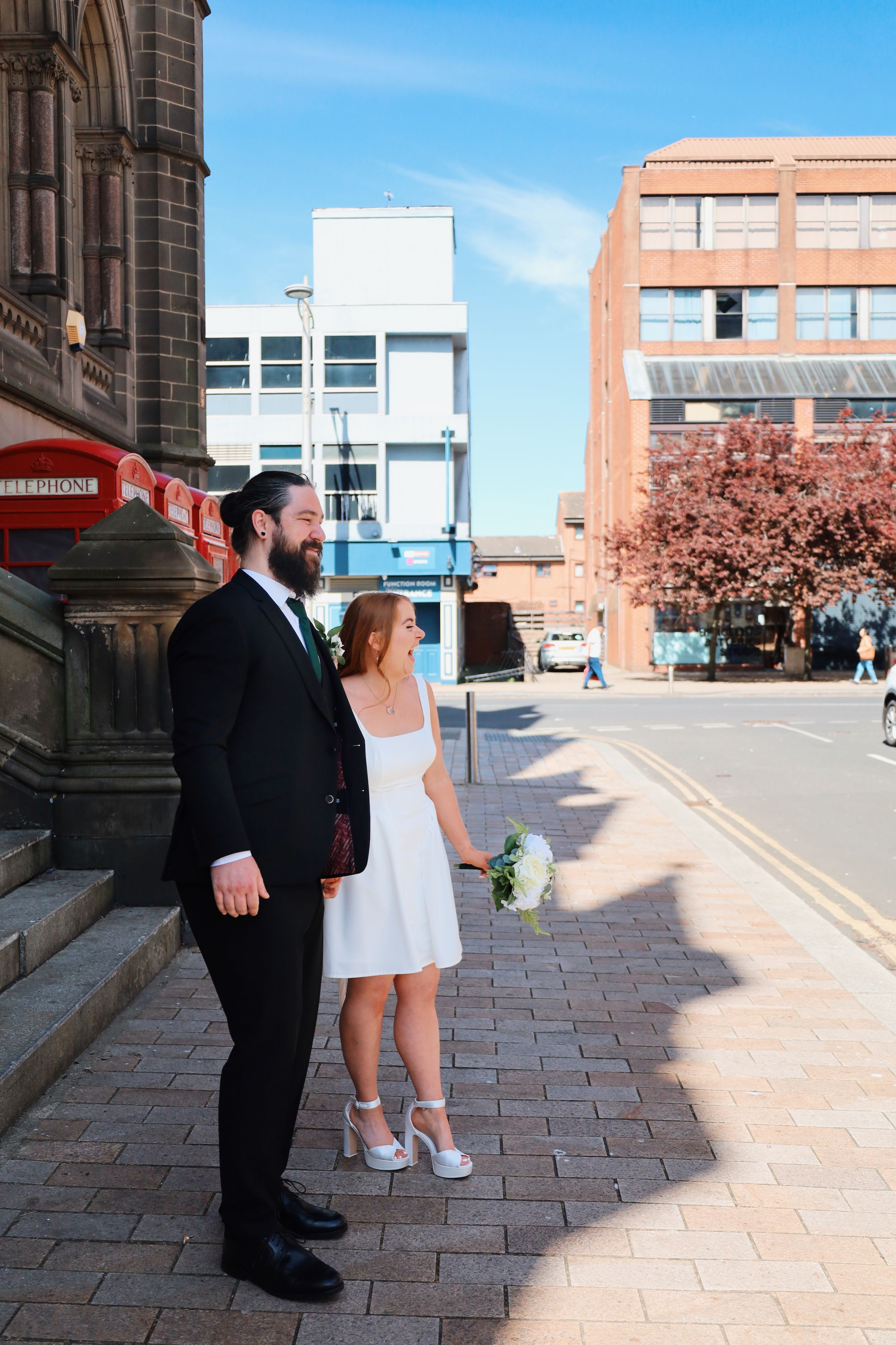 A couple poses on a stone sidewalk with modern buildings and blue sky in the background.
