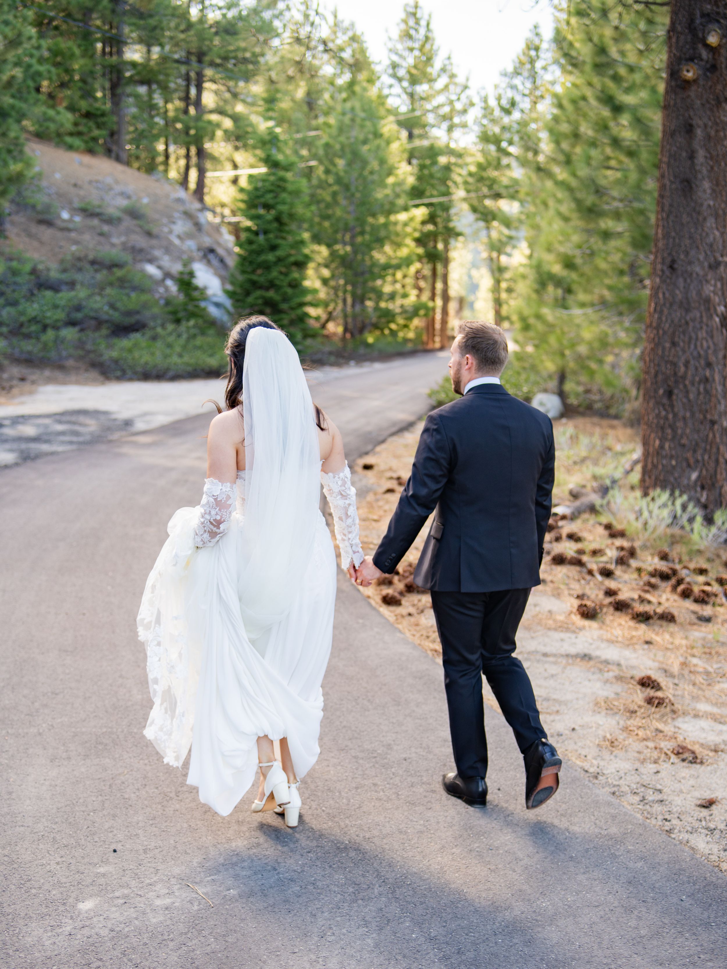 A couple walks hand in hand down a winding forest path, the bride's white dress and veil flowing behind her.