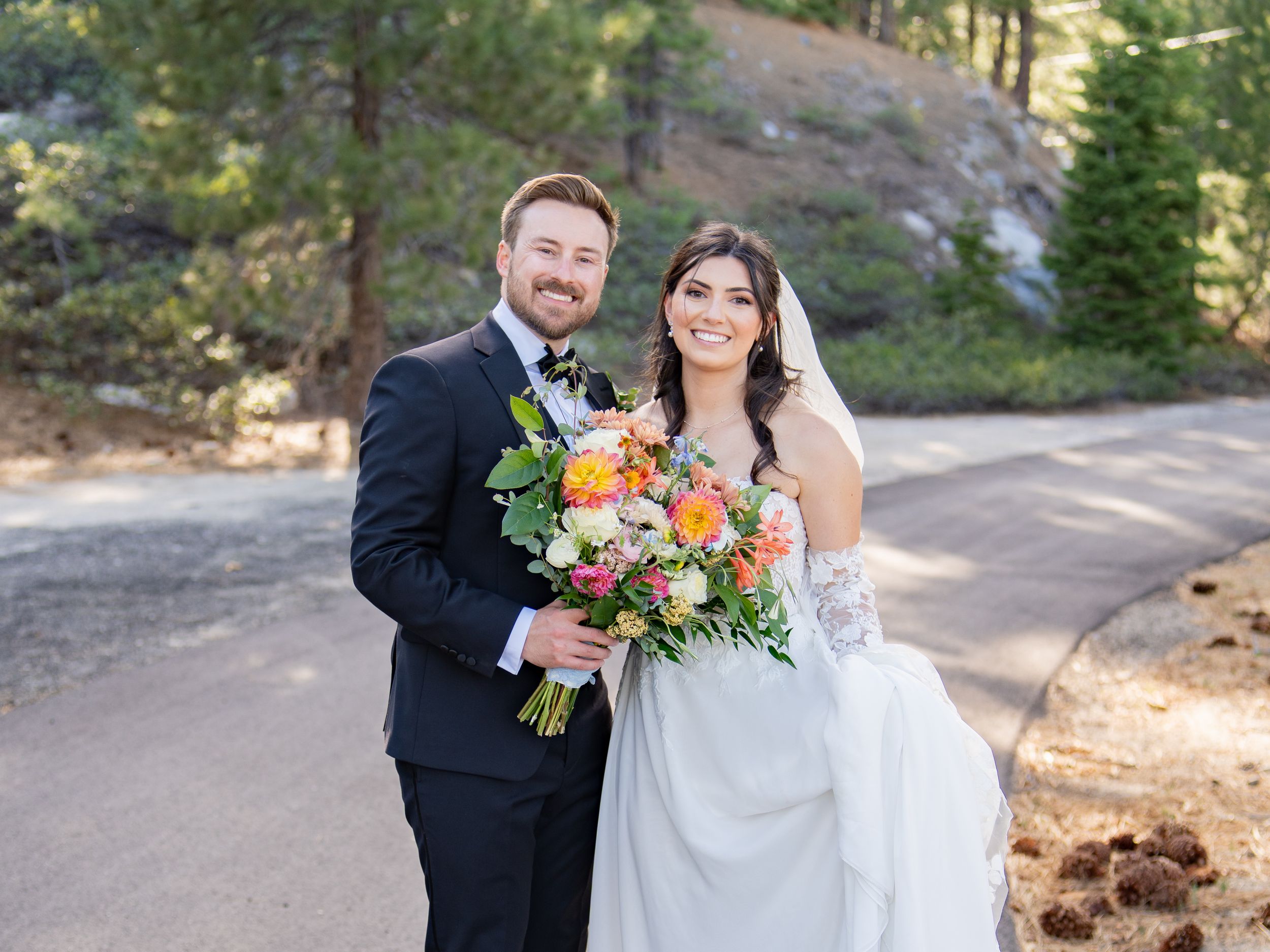 A couple in wedding attire stands together outdoors in a sunny forest setting with natural decor and beautiful lighting.
