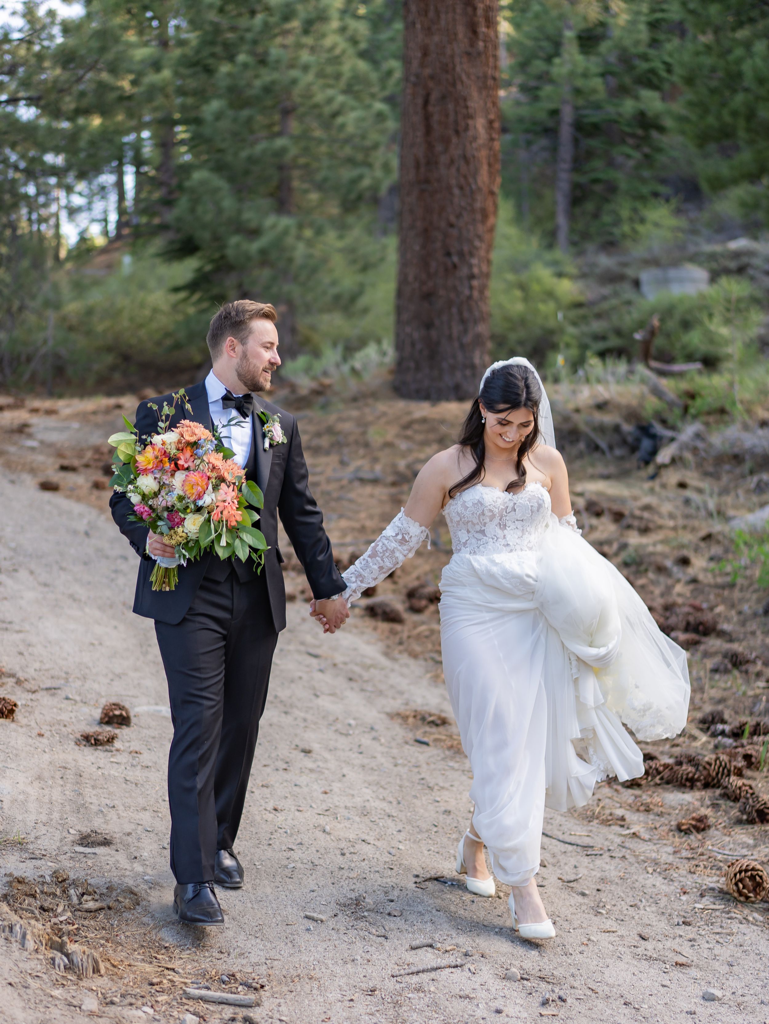 A couple in wedding attire dances playfully across a scenic outdoor trail surrounded by pine trees and rocky terrain.