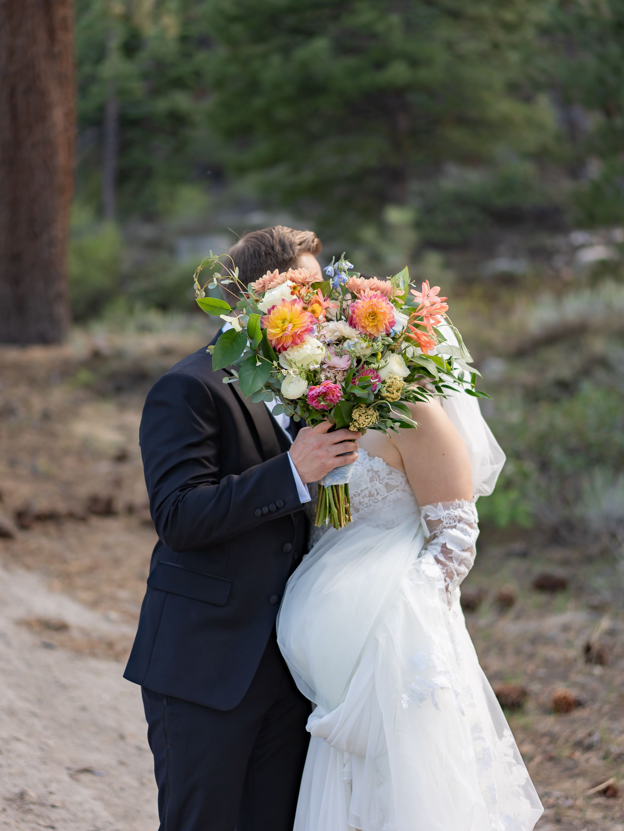 A romantic wedding moment on a forest path with a lush peach and white floral bouquet hiding behind the couple.