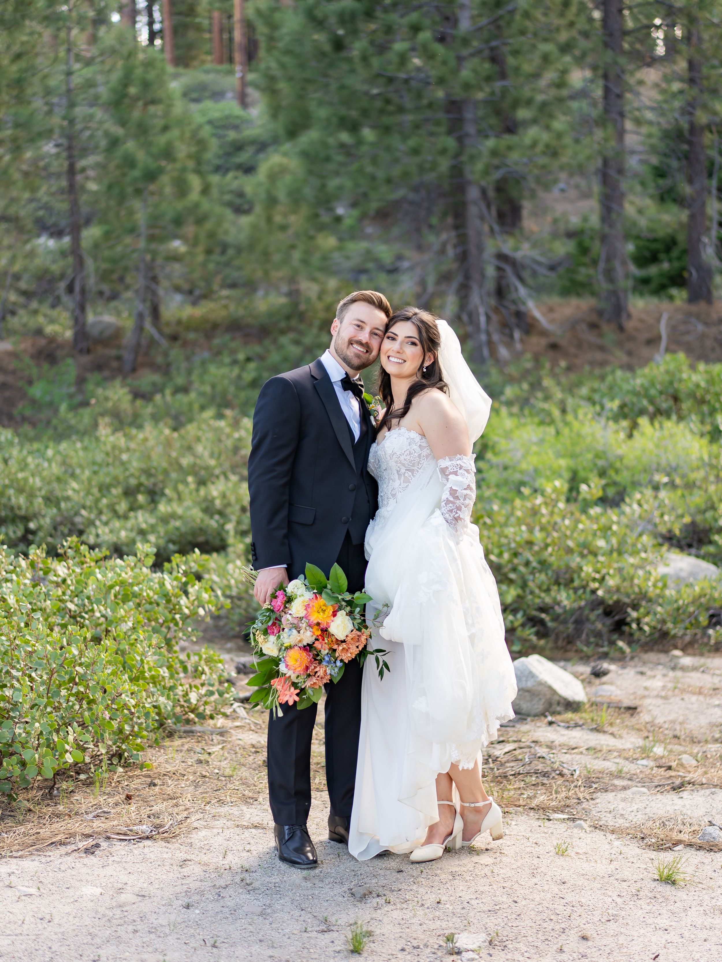 A couple poses together in a wooded forest setting with green pine trees and natural scenery in the background.