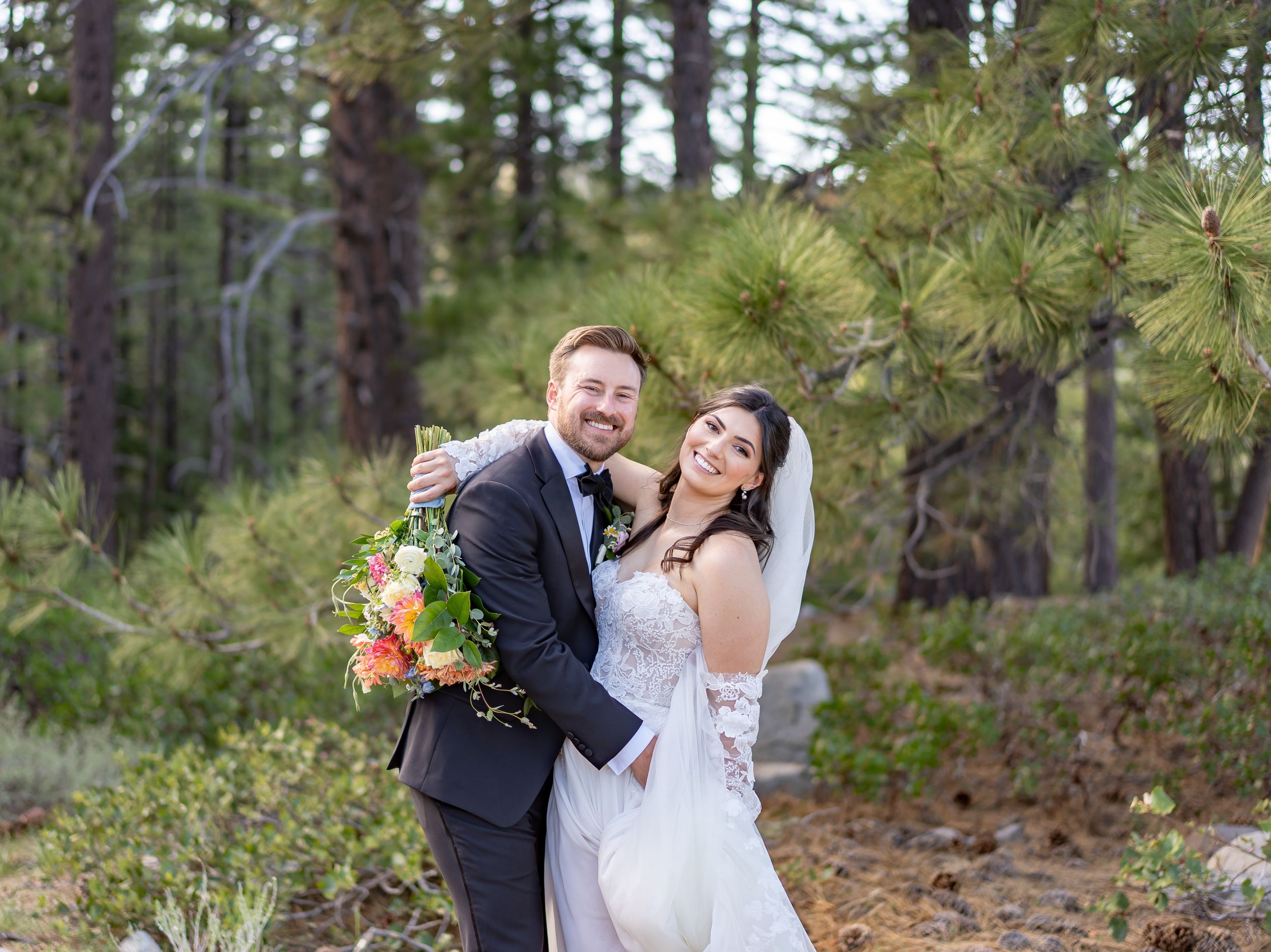 A newlywed couple shares a joyful moment in a forest setting surrounded by pine trees.