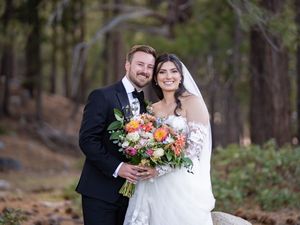 Newlyweds stand together outdoors in a wooded setting with bride holding vibrant orange and white floral bouquet.