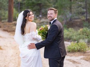 A couple in formal wedding attire embraces while walking on a wooded path in a natural outdoor setting.