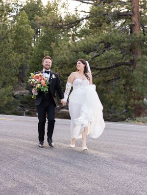 A wedding couple runs playfully along a mountain road surrounded by pine trees as the bride's dress flows in the wind.