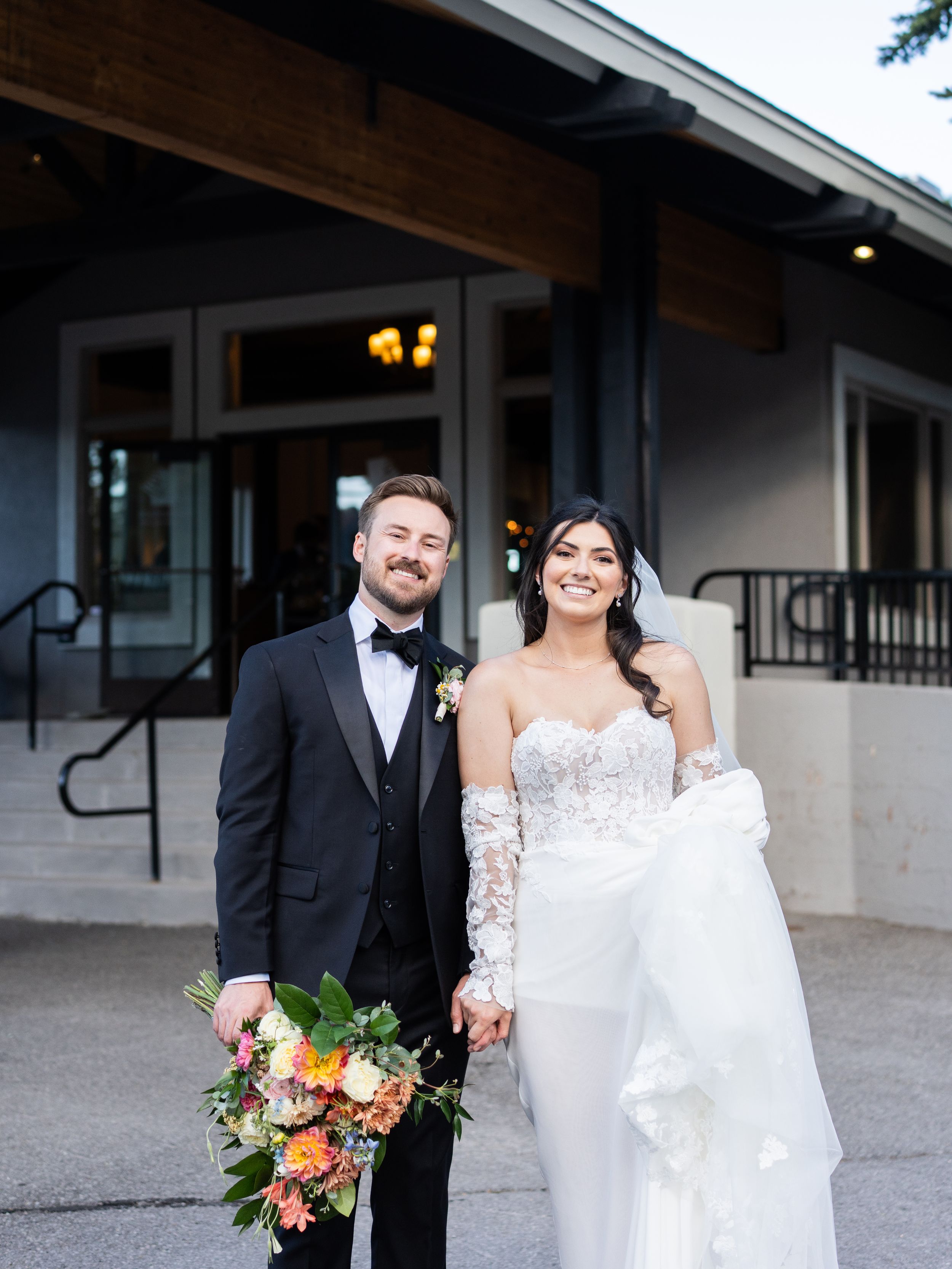 A wedding couple stands outside a rustic building entrance with a vibrant autumn-colored bouquet.