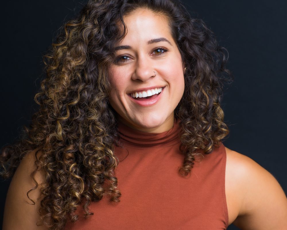 A person with long curly dark hair and a bright smile wearing a rust-colored sleeveless top against a dark background.