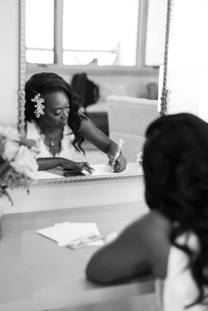 A black and white photo of a person writing at a table with a floral hair accessory seen in reflection.