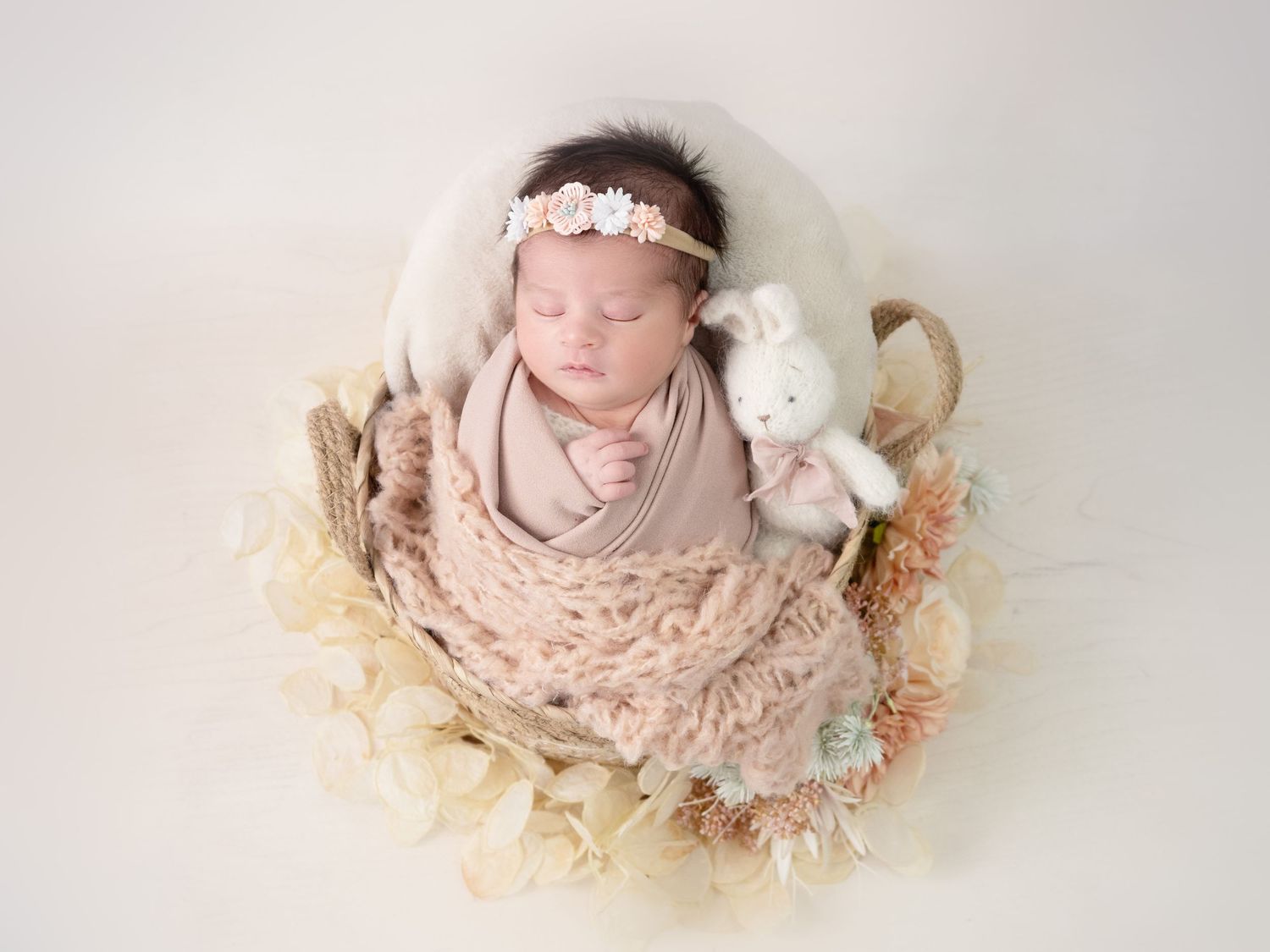 Newborn baby girl sleeps peacefully in a basket, wrapped in soft pastel pinks and creams - a dreamy, gentle moment full of warmth and love
