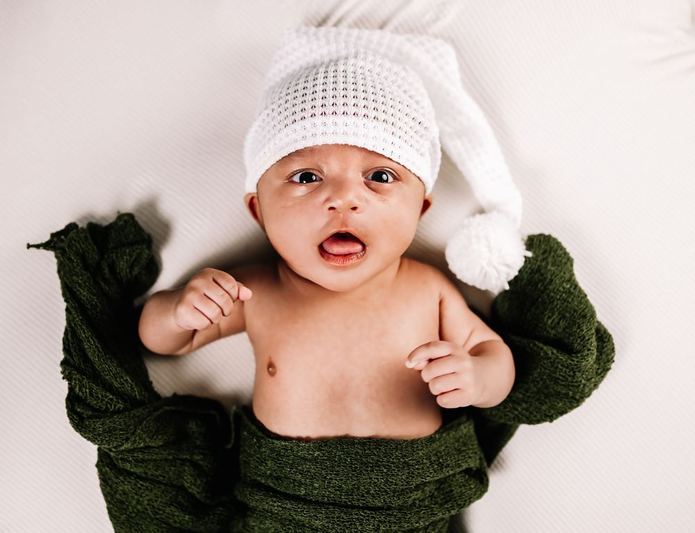Baby wearing white knit hat and green sweater lies on white background with surprised expression.