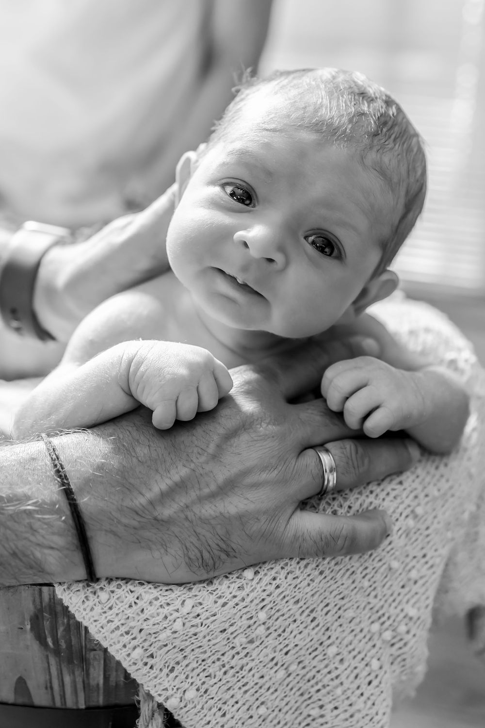 Black and white portrait of a baby lying on a textured blanket with an adult's hand visible underneath.