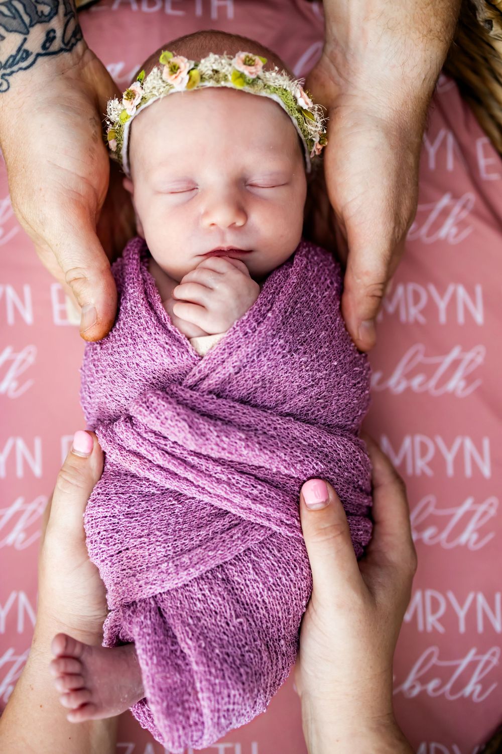 A sleeping newborn wrapped in purple swaddle with floral crown lies on pink background with text pattern.