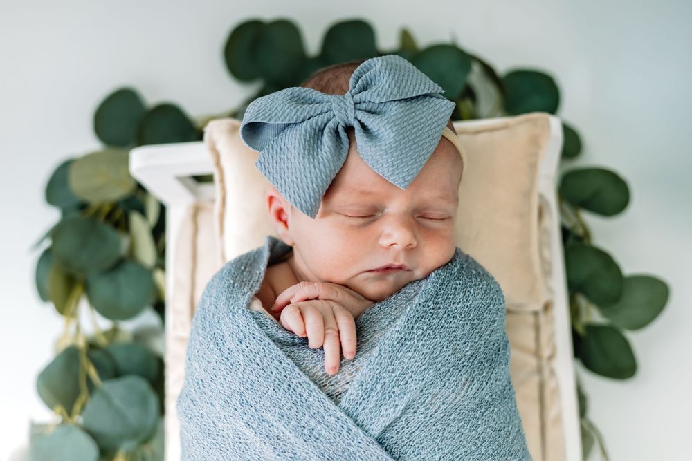 Newborn baby wrapped in soft blue swaddle blanket with eucalyptus leaves and wooden decor in background.