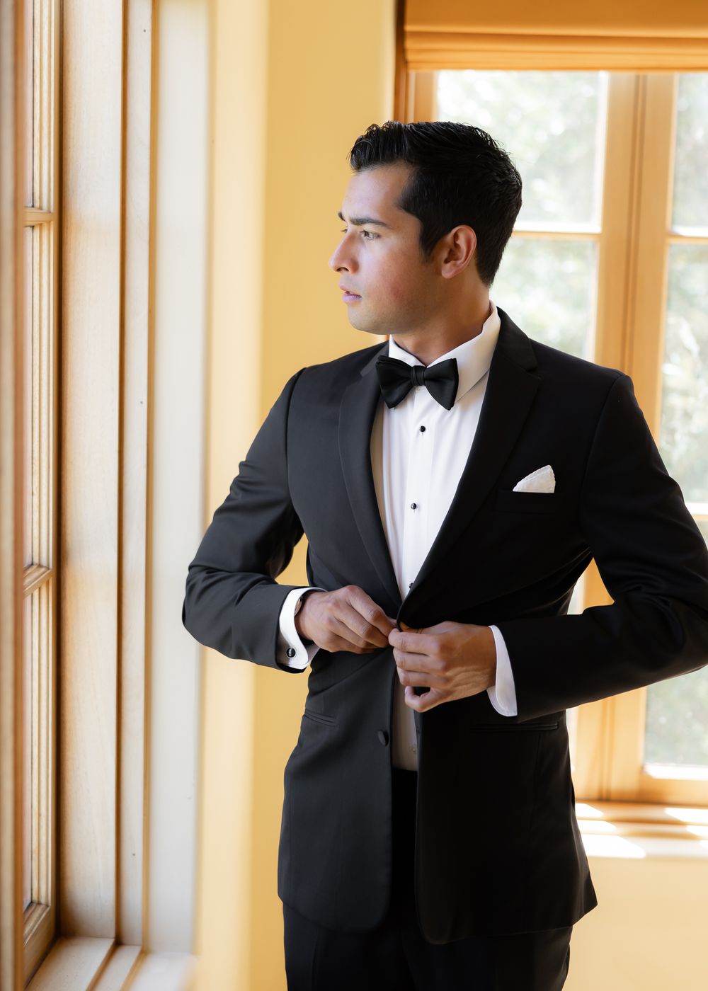 Groomsmen in black tuxedos getting ready before wedding ceremony in sunlit room with large windows.