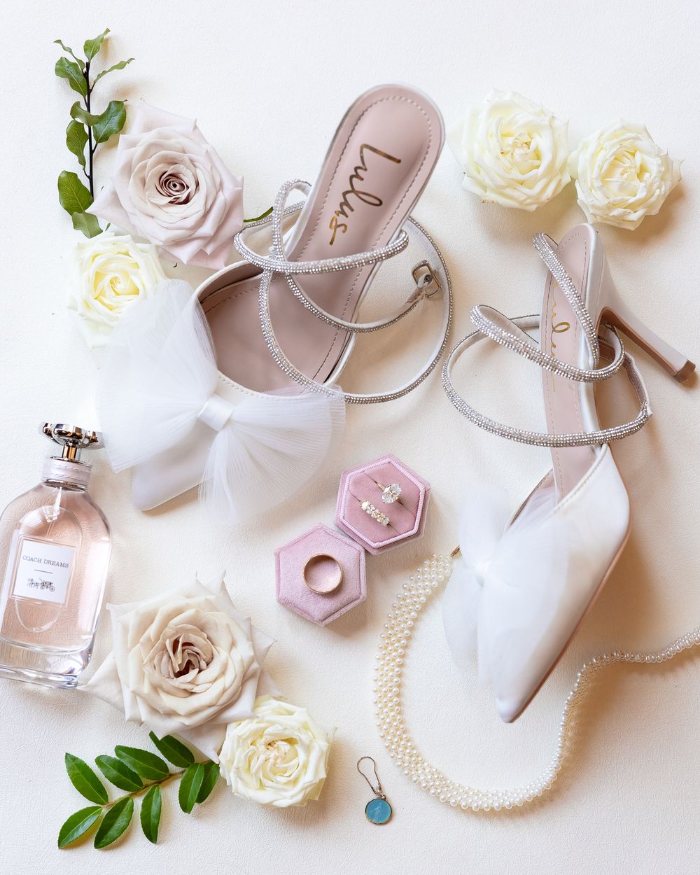 Bridal accessories including blush pink shoes, pearls, perfume bottles and white roses arranged on white background.