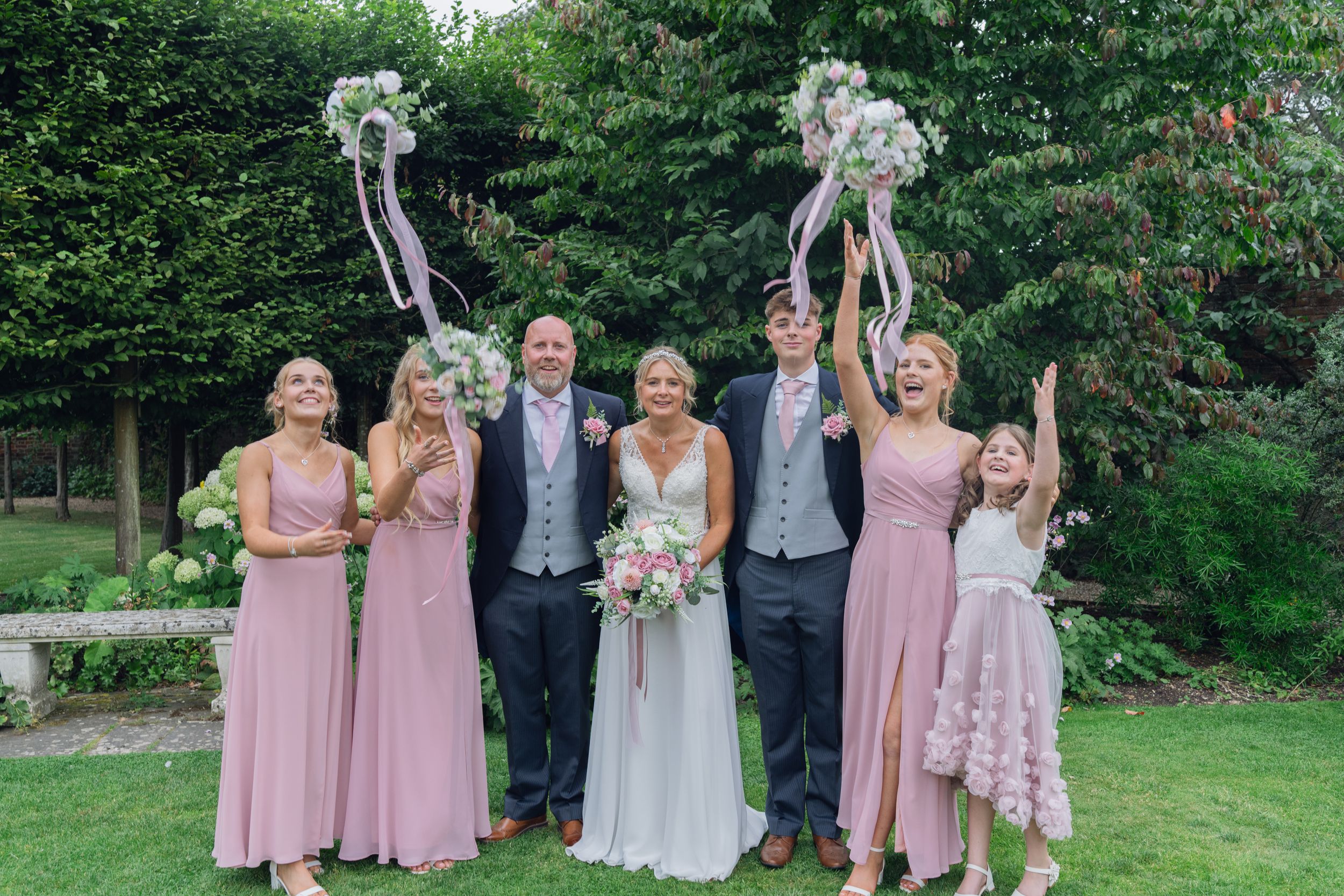 Wedding party in pink bridesmaids dresses poses together on green lawn during outdoor summer celebration.