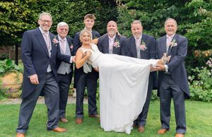 Wedding party members in navy suits and brown shoes playfully pose while lifting a bride's dress in an outdoor garden setting.