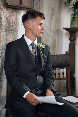 Groom in black suit with boutonniere sits holding papers while looking sideways during wedding ceremony.