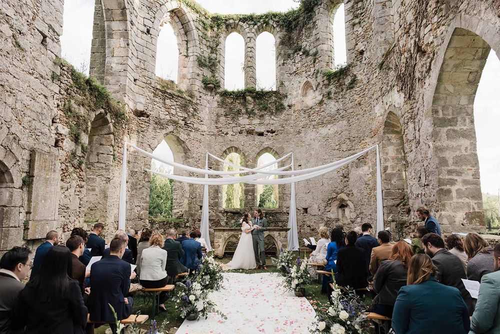 Wedding ceremony taking place in ancient stone church ruins with white fabric draped across arches.