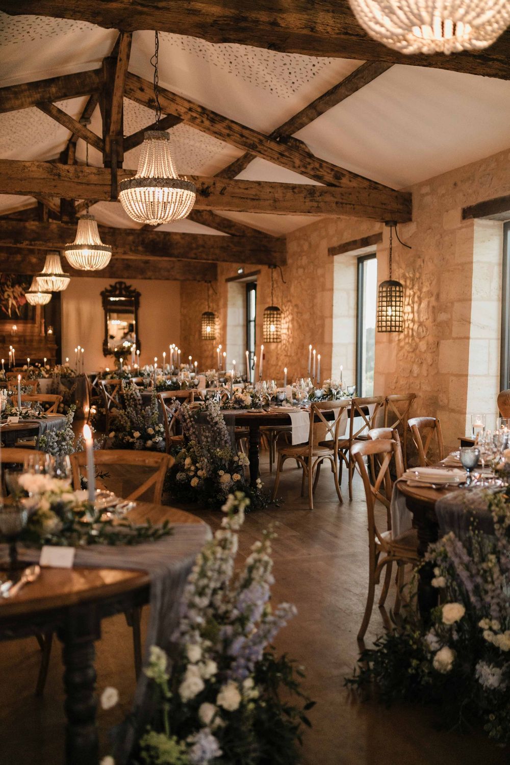 Rustic reception venue with exposed wooden beams, crystal chandeliers, and tables decorated with floral arrangements.
