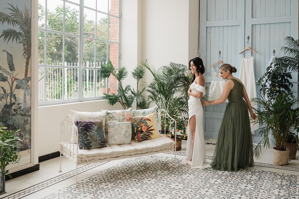 Two people stand in elegant white gowns by bright sunlit windows in a spacious room with tropical plants and tiled floors.