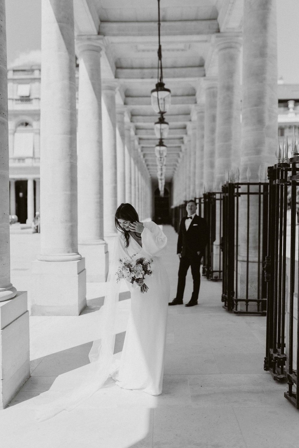 A couple shares an intimate moment in an elegant colonnade hallway filled with white pillars and hanging lanterns.