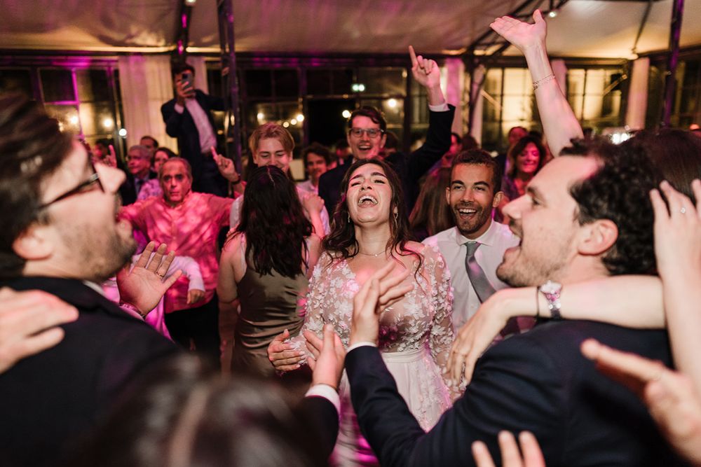 A crowd of party guests dancing and laughing together on a dimly lit dance floor with pink lighting.