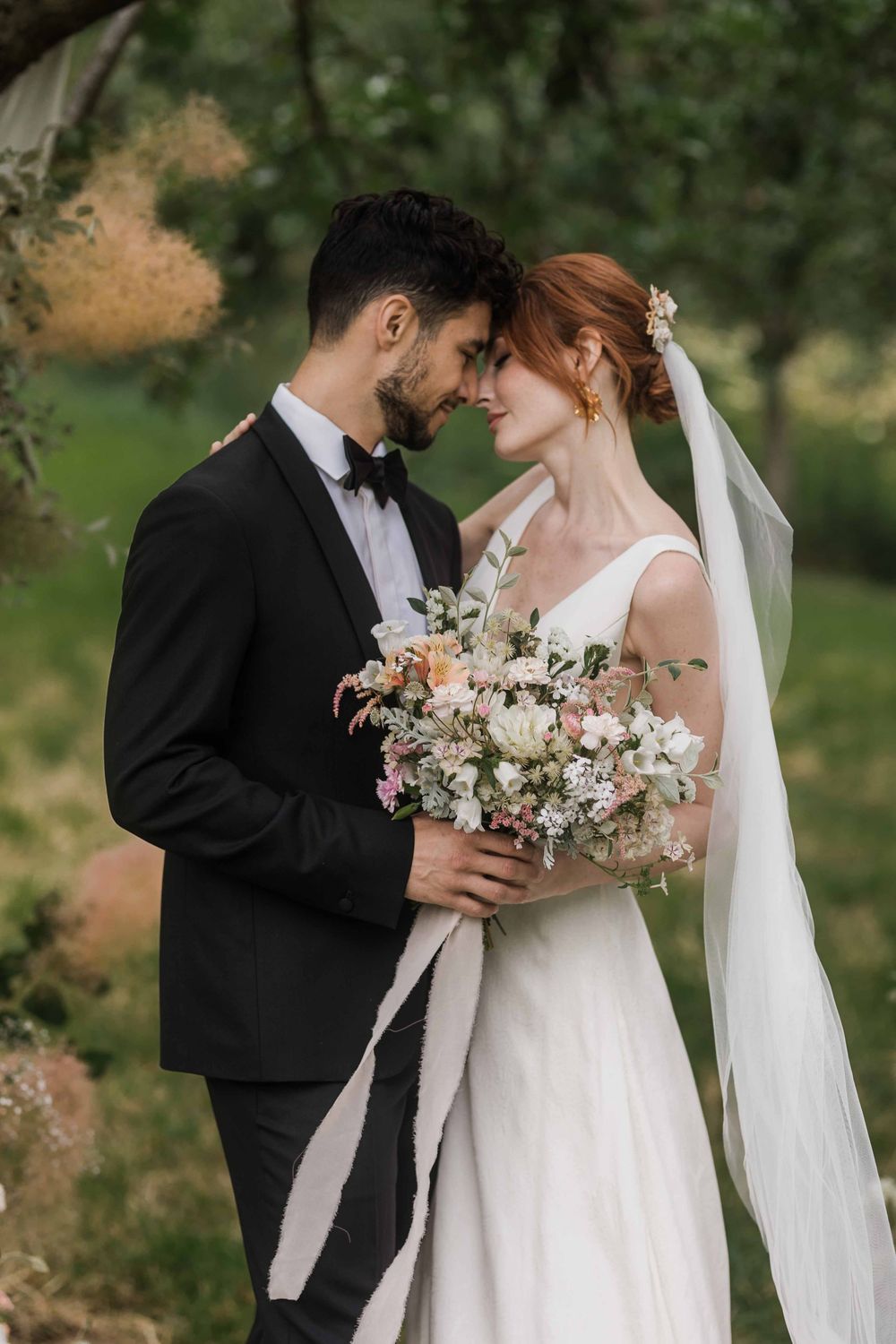 Romantic newlyweds share intimate moment in garden setting with bride holding lush pastel floral bouquet.