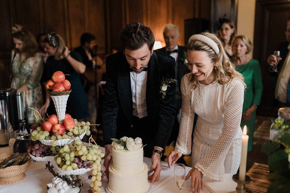 A couple cuts their multi-tiered white wedding cake together at their reception while guests look on in the background.