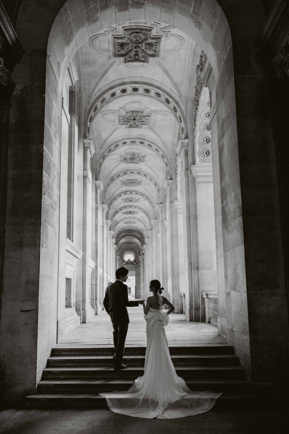 A couple stands in an elegant arched corridor with ornate vaulted ceilings and classical architecture in black and white.