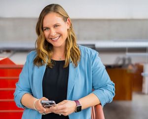 Professional wearing a light blue blazer smiles while seated at a desk using a mobile phone in an office setting.