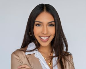 Professional headshot of a young woman with long dark hair wearing a beige blazer and white blouse, smiling at camera.