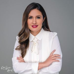 Professional woman in white blouse with bow, arms crossed, smiling confidently against gray background.