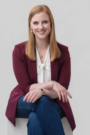 Smiling redhead woman in burgundy blazer and jeans sits relaxed against gray background.