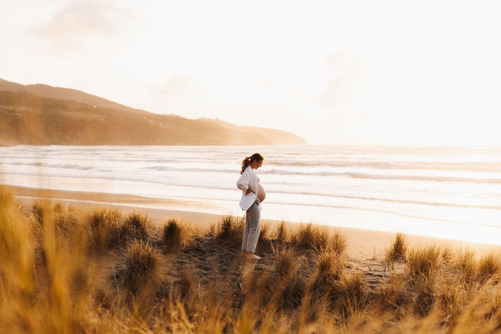 Person standing on grassy coastal dunes at sunset overlooking a serene beach with golden light in the background.