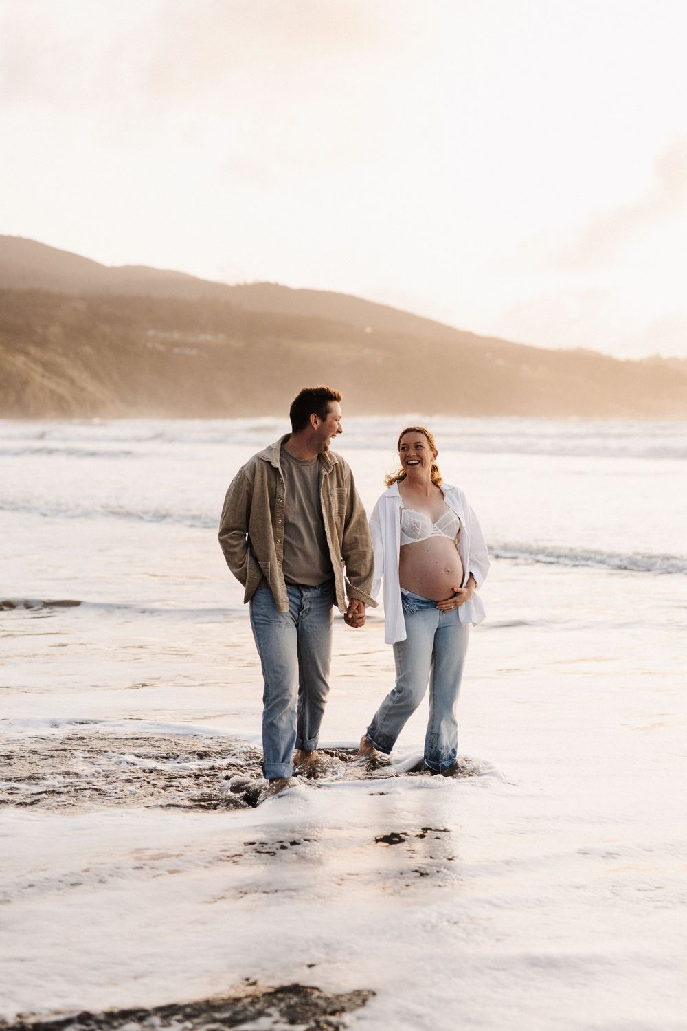 A couple walks hand in hand along a misty beach at sunset with mountains in the background.