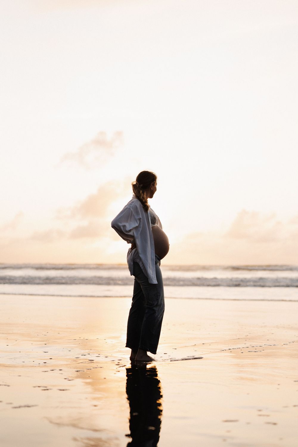 Silhouette of an expectant mother standing on a sandy beach at sunset with waves in the background.