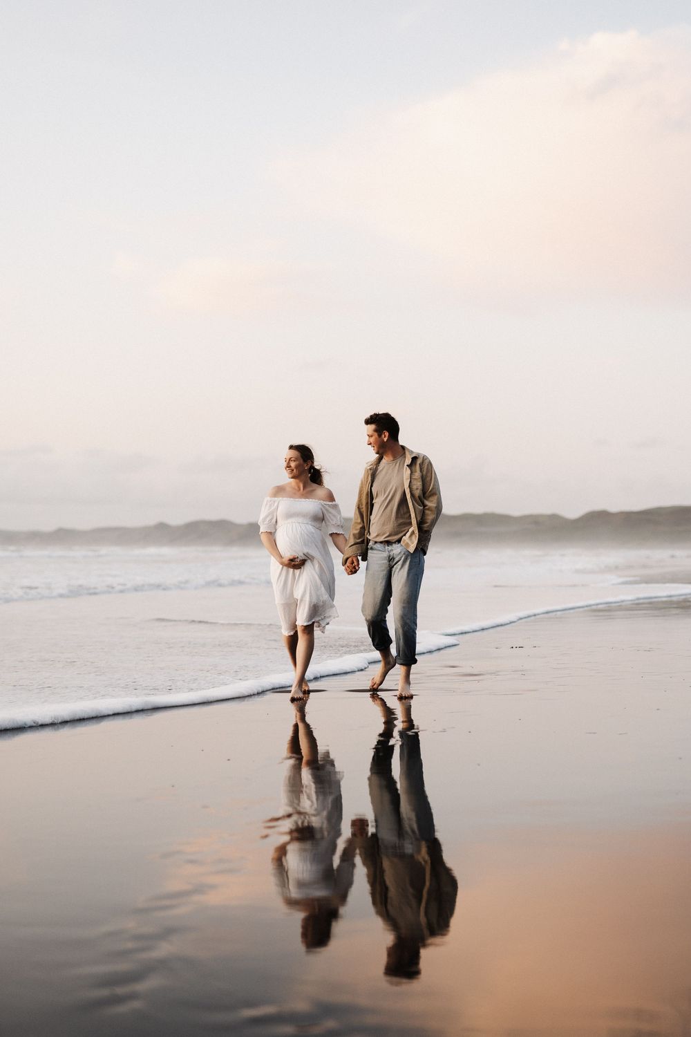 Two people walking on a beach at sunset with their reflections mirrored in the wet sand along the shoreline.