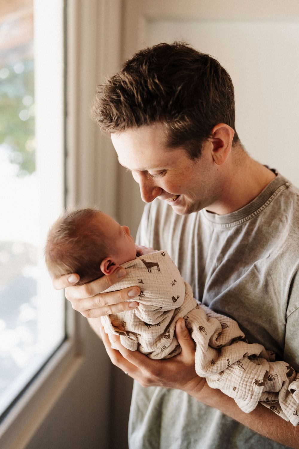 A person in a gray sweater tenderly holds a sleeping newborn wrapped in a patterned knit blanket by a bright window.