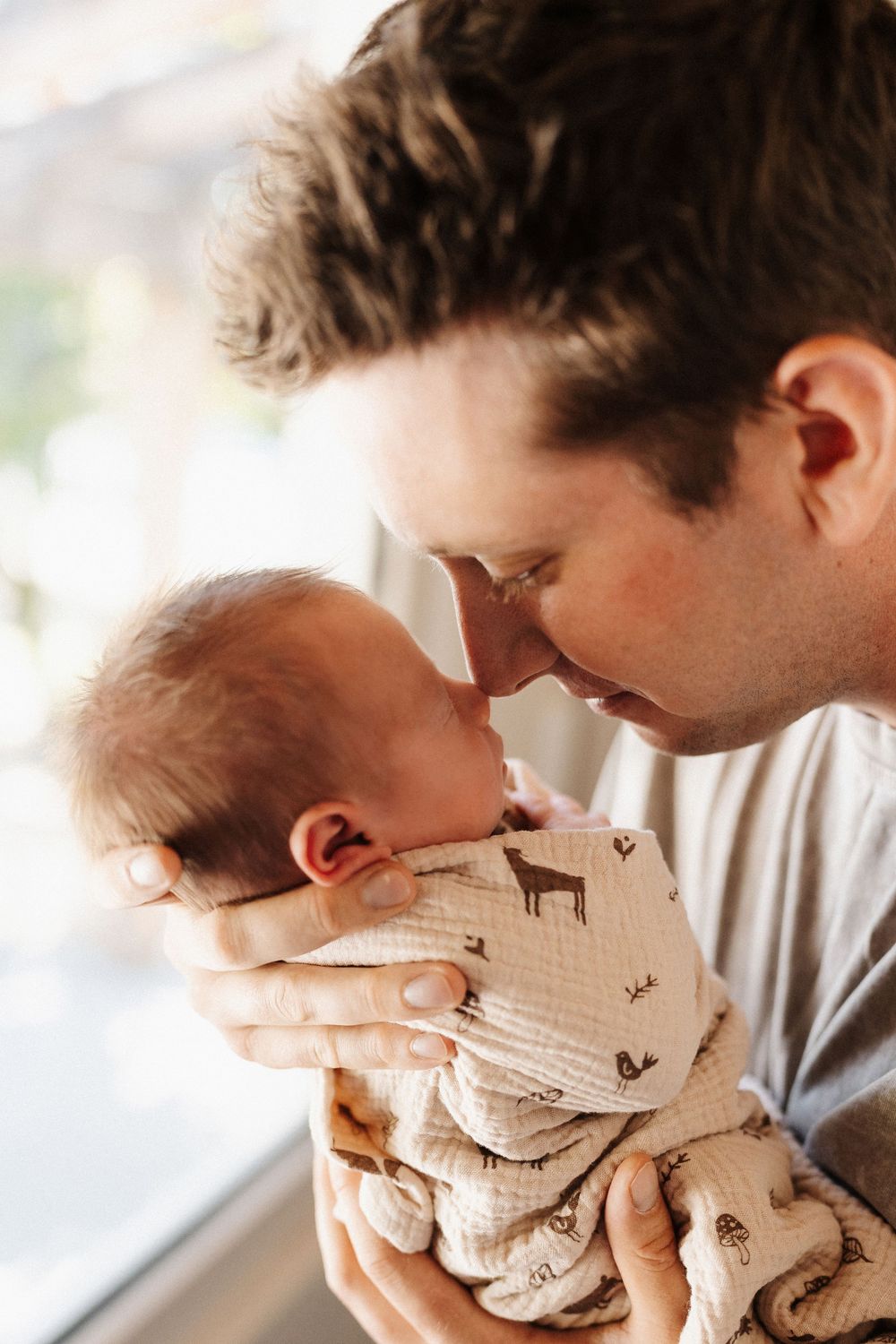 A parent tenderly holds and kisses a swaddled newborn baby against a bright window background.