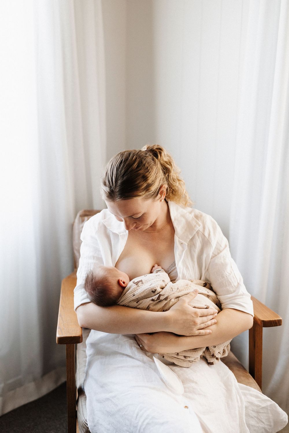 A person in white clothing sits in a chair by a window while nursing a baby in a bright, minimalist setting.