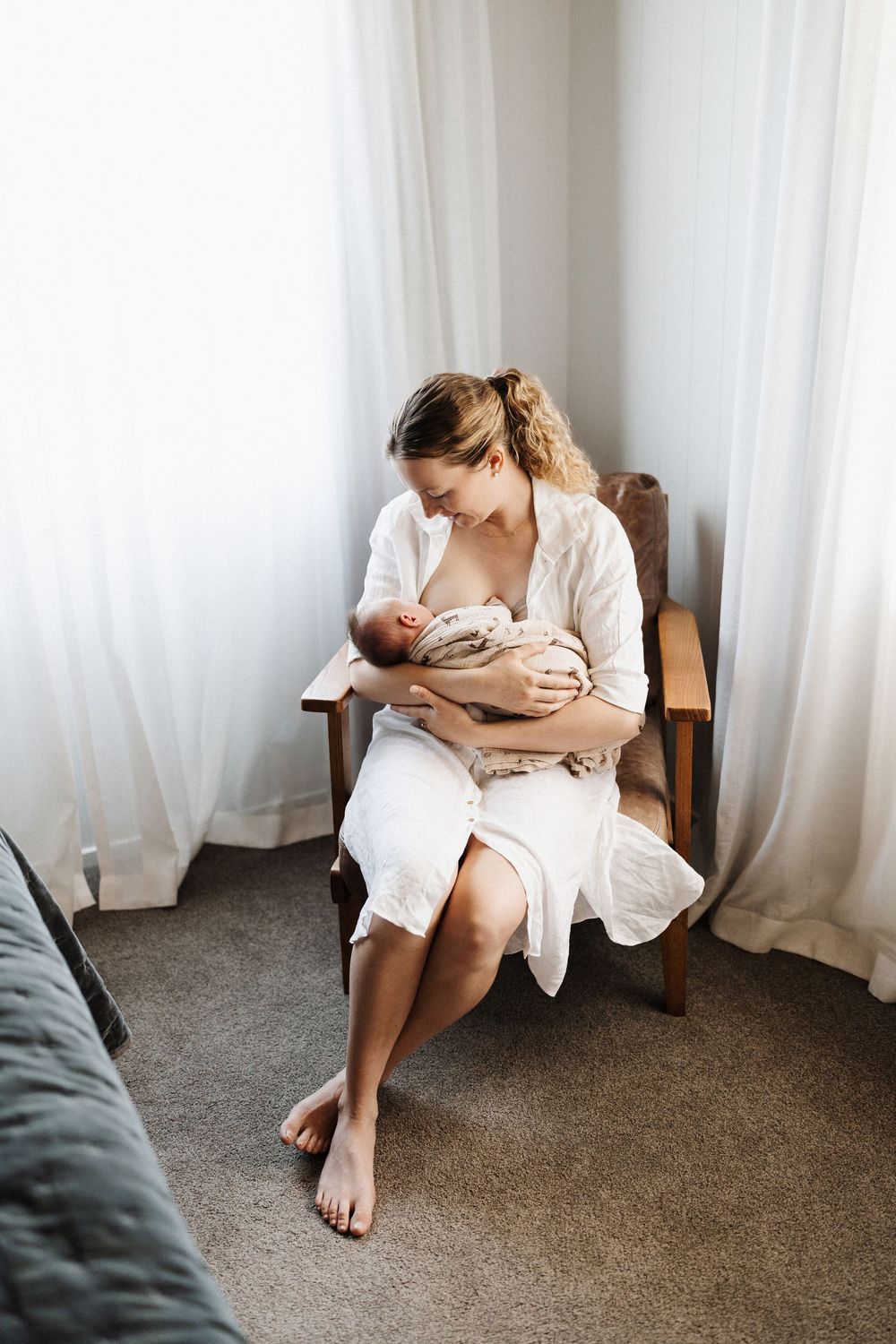 A tender moment of mother cradling a newborn baby in a white robe while sitting in a wooden chair by sheer curtains.