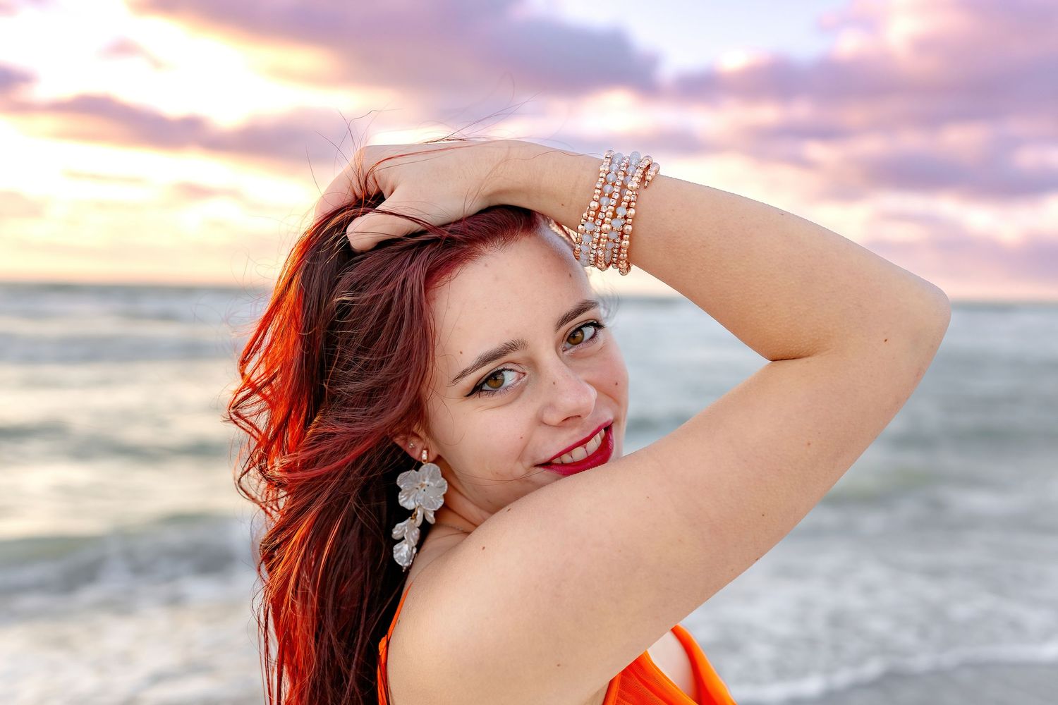 Woman in orange swimwear posing at sunset on a beach with ocean waves and cloudy sky in background.