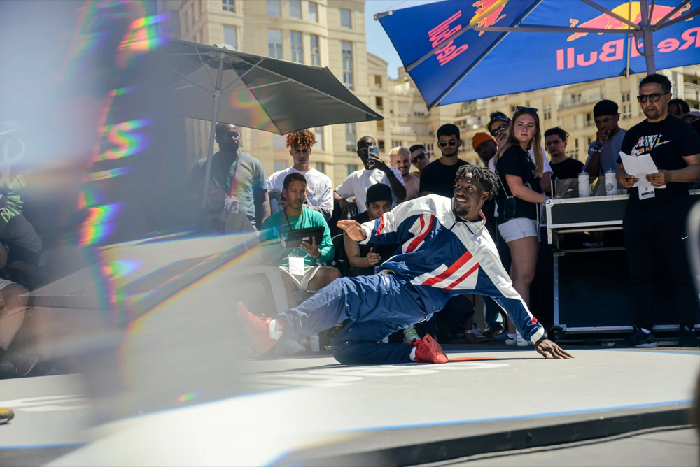 Scène dynamique de breakdance en extérieur, un danseur effectue un mouvement au sol devant le public.